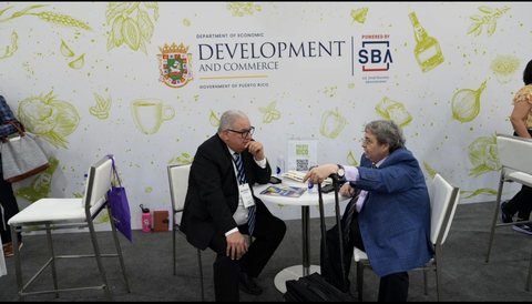Two business participants in conversation at a meeting table in front of a Puerto Rico economic development backdrop.