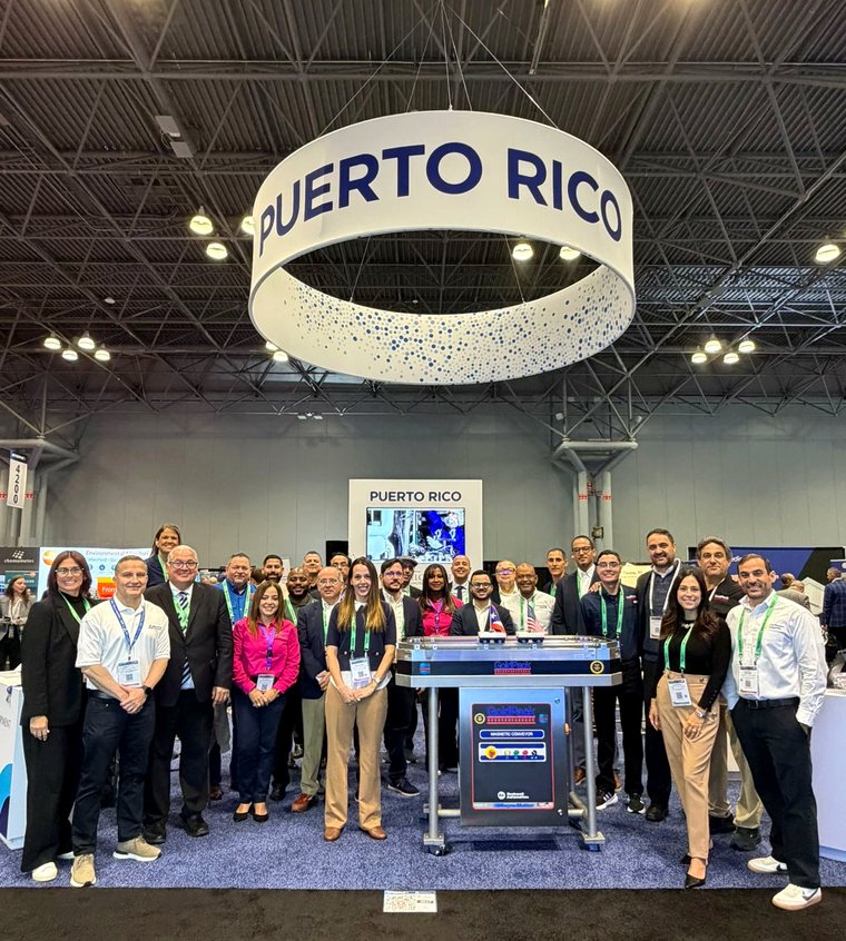 A large group of business delegates and organizers standing together beneath a circular PUERTO RICO sign at an exhibition stand.